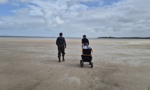 Two researchers walk a cart of sampling gear across wide sand flats beside the shallow water of the Coorong.