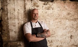 Chef Fabian Lehmann in a white shirt and black apron, arms folded, against a rough limestone wall