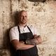 Chef Fabian Lehmann in a white shirt and black apron, arms folded, against a rough limestone wall