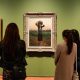 Gallery visitors standing in front of three framed paintings hung on a green wall