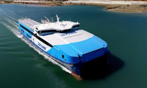 A large blue and white SeaLink ferry seen from above, moving through calm water near a sandy shoreline.