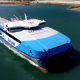 A large blue and white SeaLink ferry seen from above, moving through calm water near a sandy shoreline.