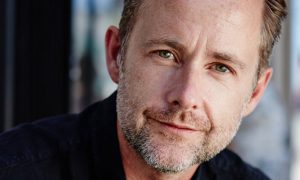 Actor Billy Boyd in a dark shirt, smiling beside a sunlit window